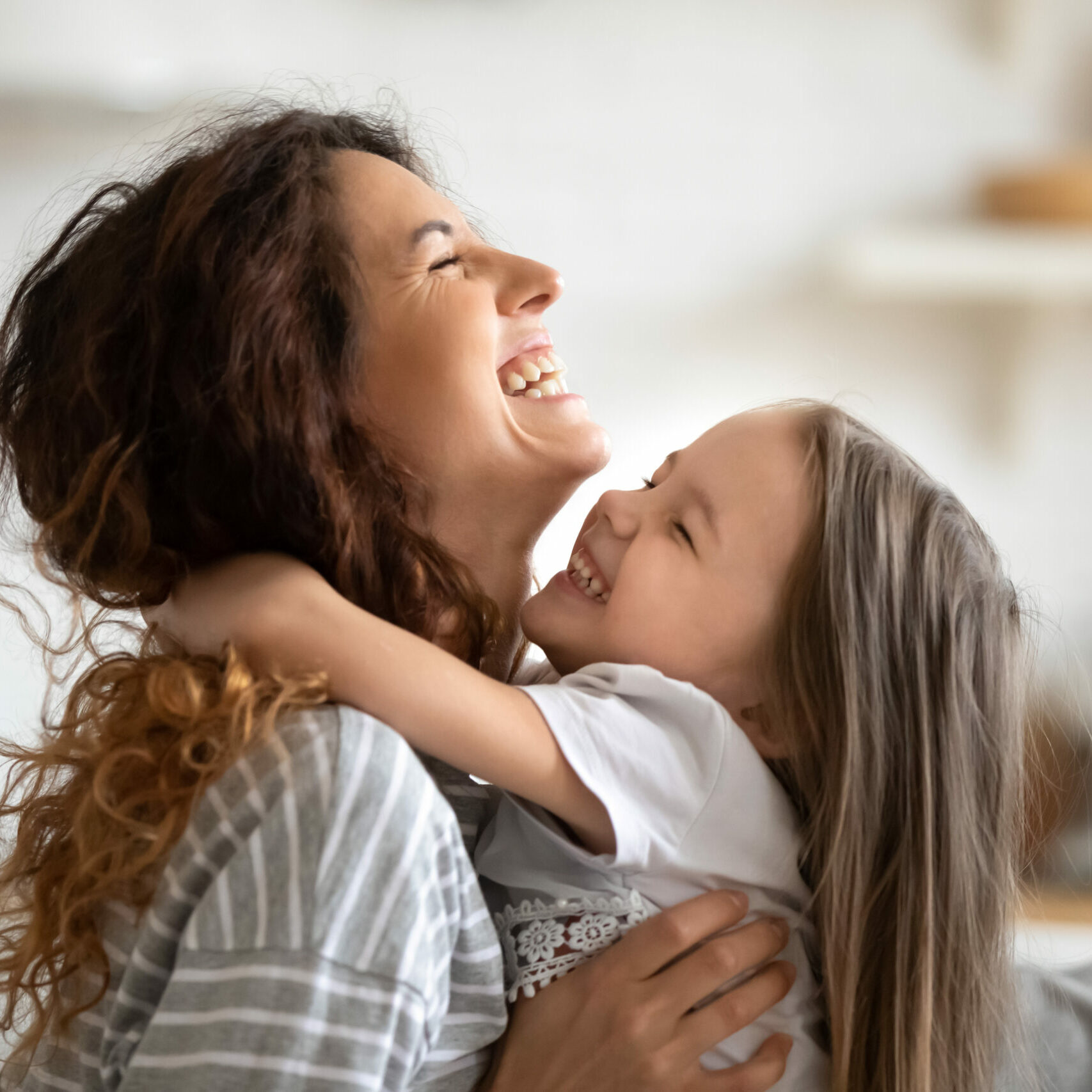 Close up side view overjoyed smiling young mother and daughter hugging and laughing, enjoying tender moment, happy mum and adorable preschool girl kid cuddling, having fun together at home